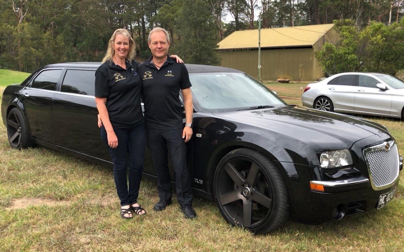 A photo of Darren and Julia, the owners of Elude, and escape room enthusiasts, both happy while standing in front of a stretched black Chrysler 300